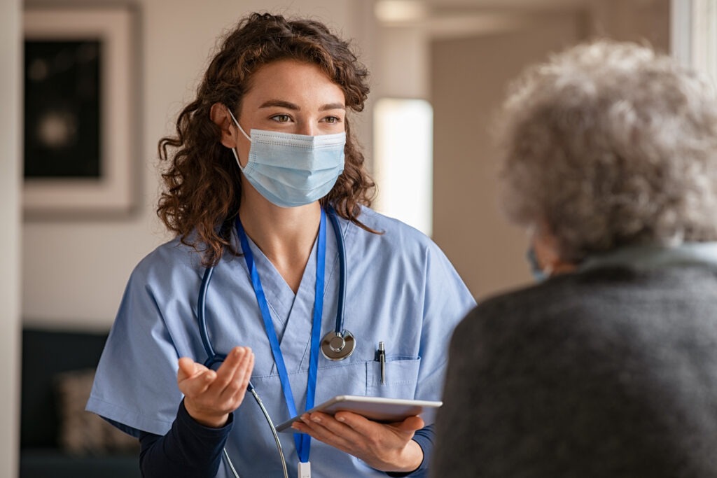 Doctor wearing safety protective mask supporting and cheering up senior patient during home visit during covid-19 pandemic.